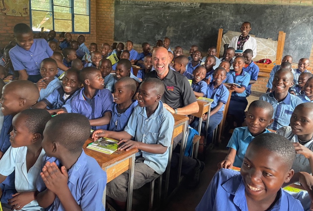 A school class with smiling children sitting at their desks. Christopher Miles sits in one seat and smiles.